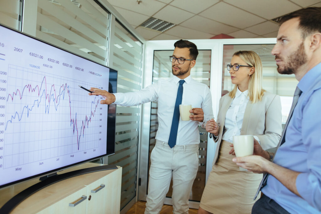 Businessman with coffee presenting growth chart on TV monitor to two co-workers.