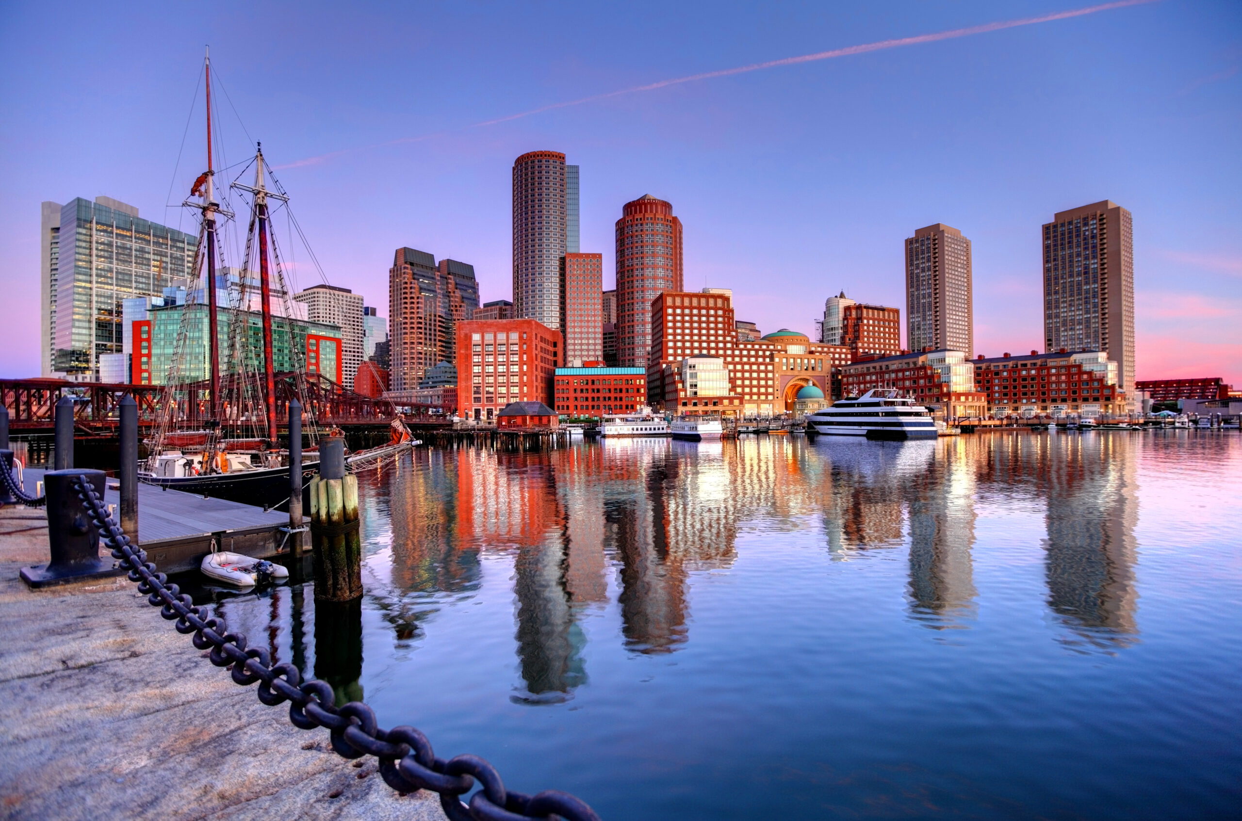 Boston skyline at dusk with reflections in the water, featuring historic ships and modern buildings under a colorful sky.