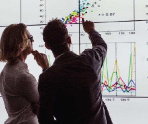 Two people analyze data on a large screen displaying various charts and graphs, including one with 'r = 0.87'.