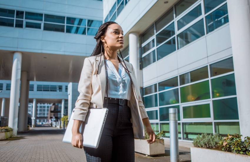 Young, female business professional standing outside of an office building, holding her laptop as she looks up at the office.