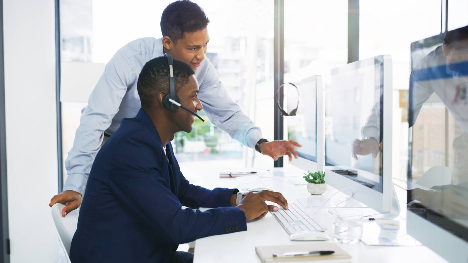 Male manager assists a call center representative who is seated at his desktop computer with a headset on.