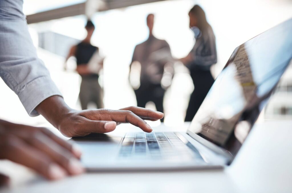 Close-up of a person's hands typing on a laptop keyboard, with blurred figures of people in the background.