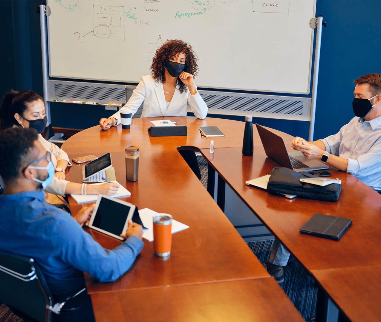 Woman leading a boardroom meeting at the head of the table with colleagues working on their devices. Covid masks