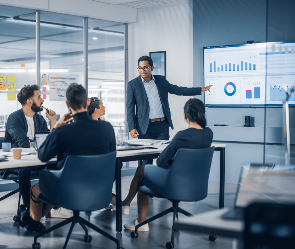 Business meeting with man presenting results on a metrics dashboard to his colleagues sitting at a boardroom table and listening.