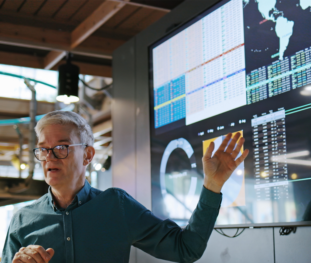 Senior businessman speaking to data being presented on TV monitor behind him