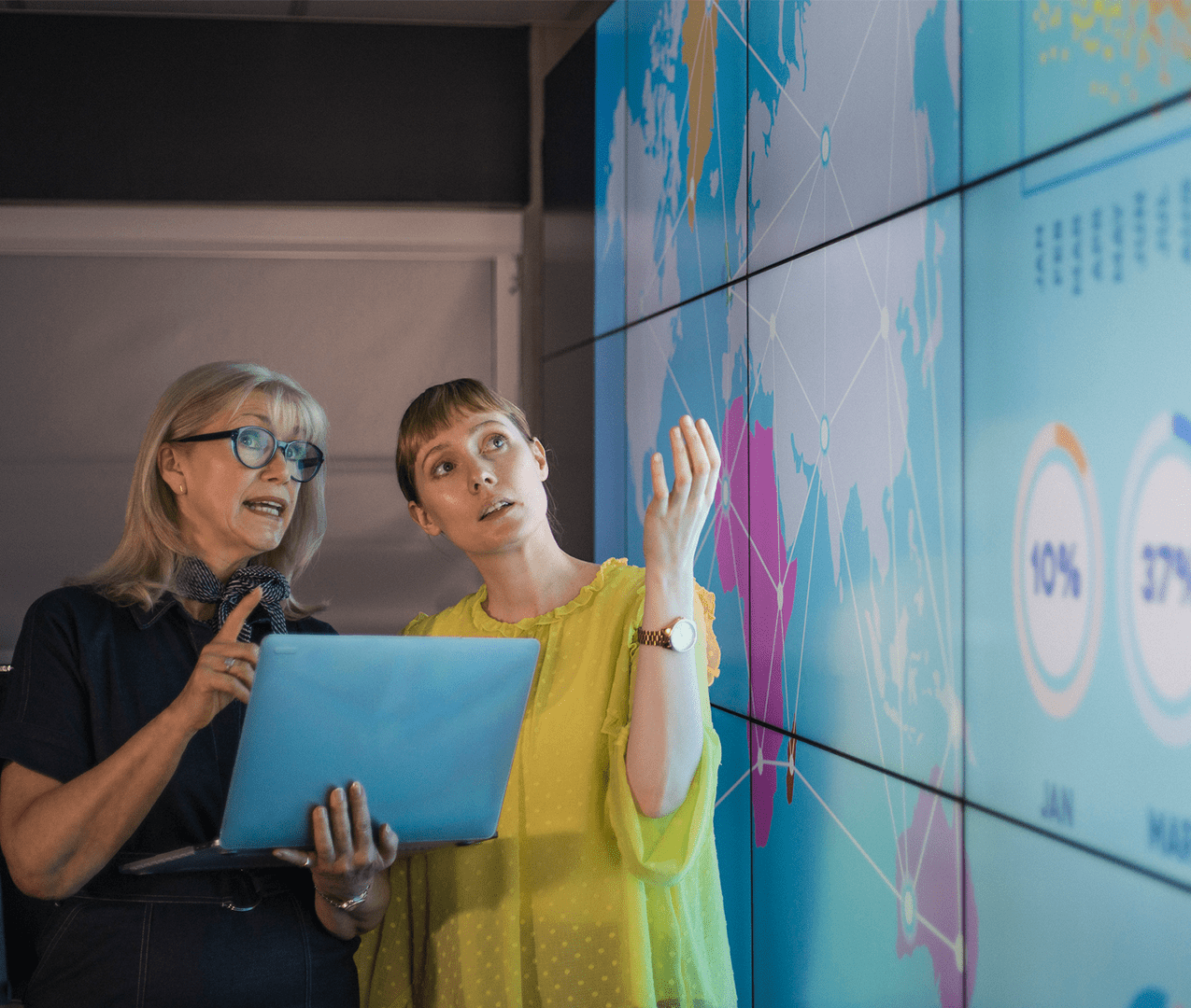 A senior business woman reviews data projected on a large screen in a boardroom with her younger colleague.