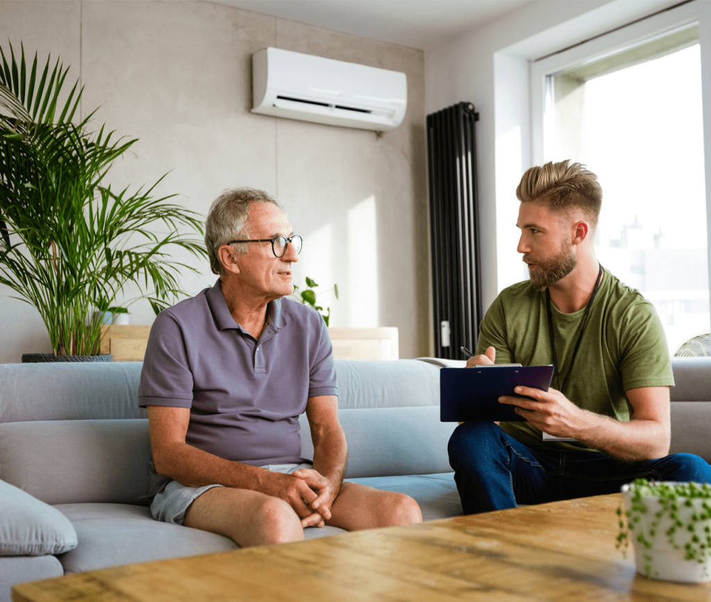 District nurse talking with senior man sitting on a couch during a home consultation. The nurse is asking the patient questions and taking notes on a clipboard.