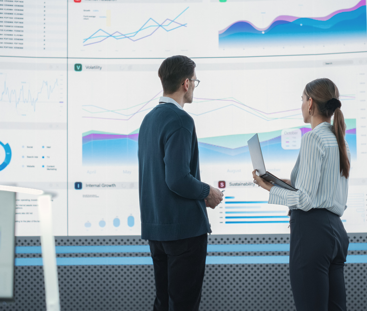 Businesswoman holding computer standing in front of a very large screen discussing the charts shown with businessman