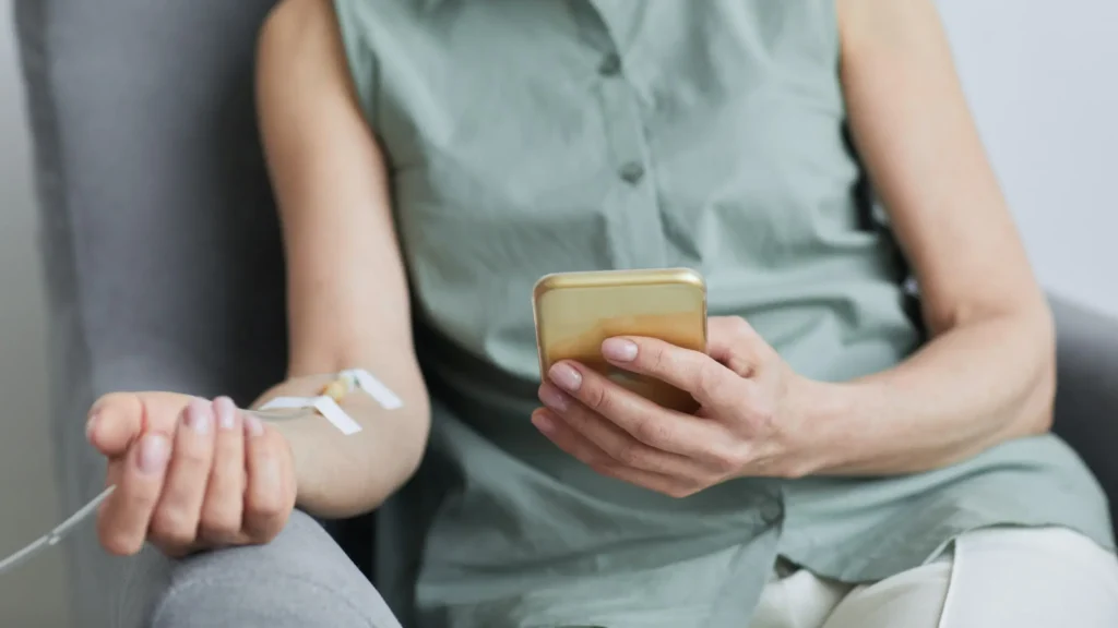 A person with an IV in their arm uses a smartphone while sitting in a chair, focusing on the technology.