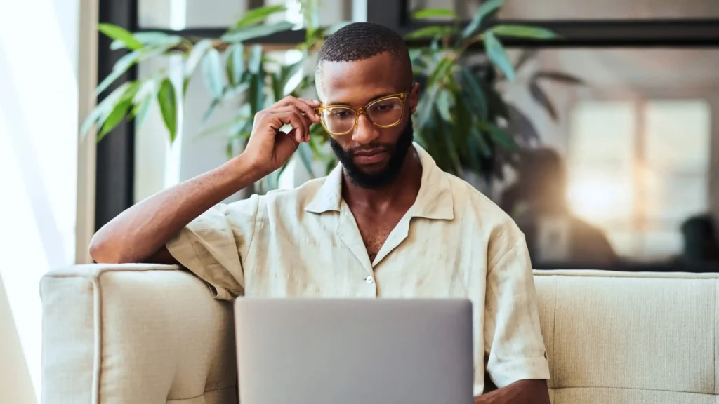 A man with glasses and a beard sits on a couch, looking down at a laptop in front of him. He is wearing a light-colored shirt.