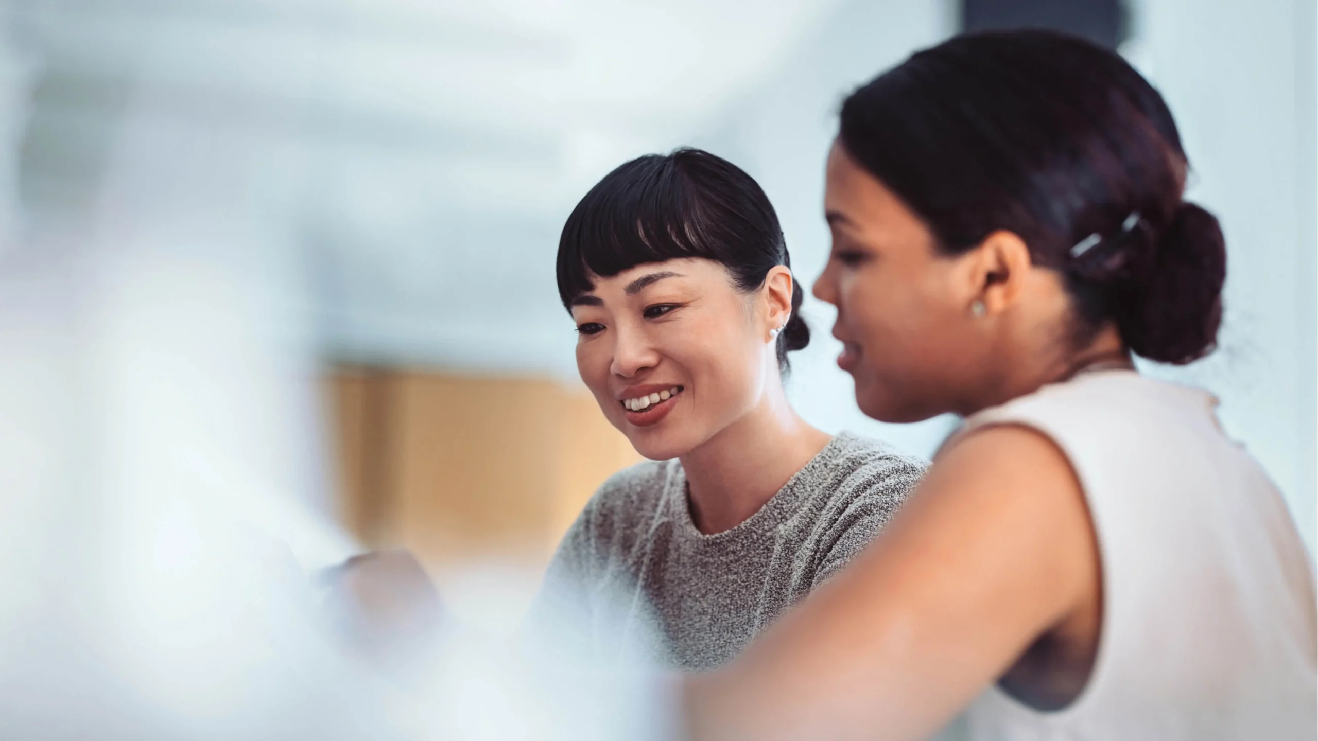 Two women in a professional setting, one smiling while looking down, suggesting collaboration or discussion.