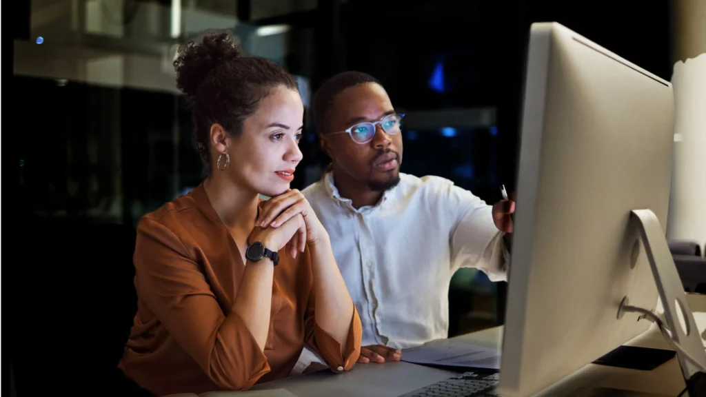 Two diverse professionals, a man and a woman, collaborate intently on a computer screen in a dimly lit office. The man points at the monitor while the woman observes.