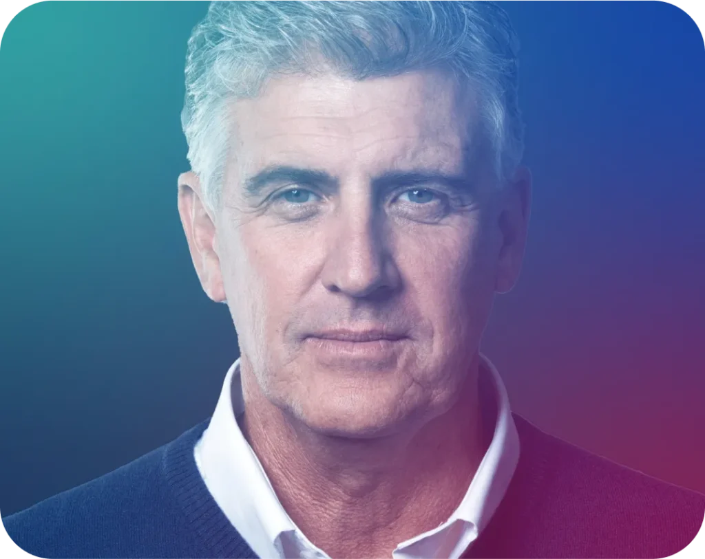 A headshot of a man with gray hair and blue eyes, wearing a white shirt and a dark blue sweater, against a gradient background.