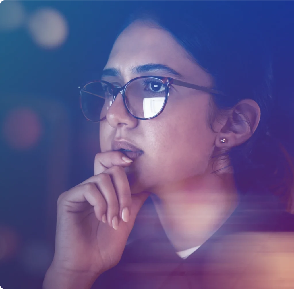 A woman in glasses looks intently at a screen, her hand near her mouth, with reflections visible in her lenses.
